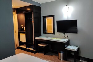 A compact hotel room with a desk, a television, a wardrobe, and a small bench.