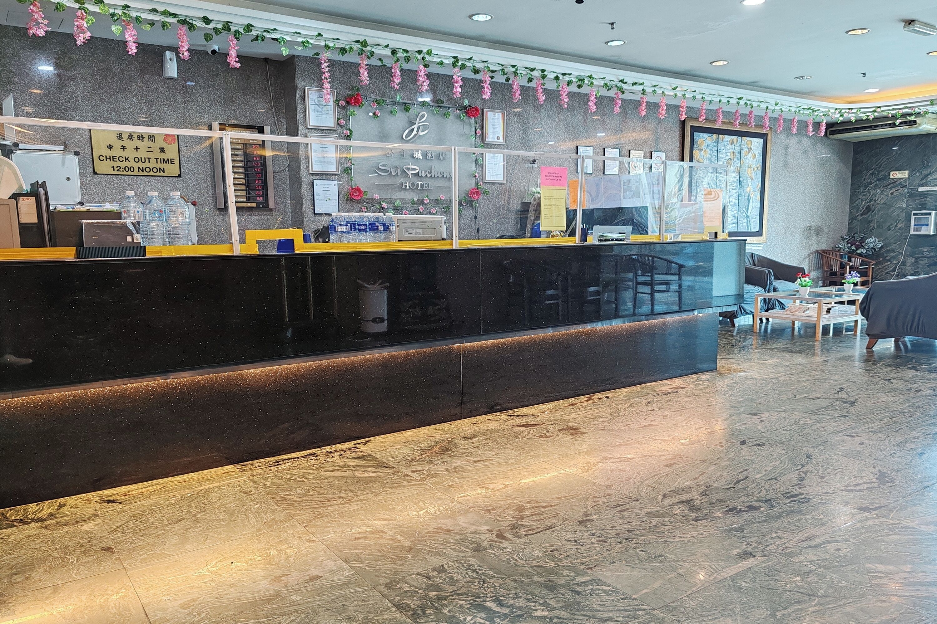 A hotel reception area with a check-out counter, a digital screen, and a seating area with tables and chairs.