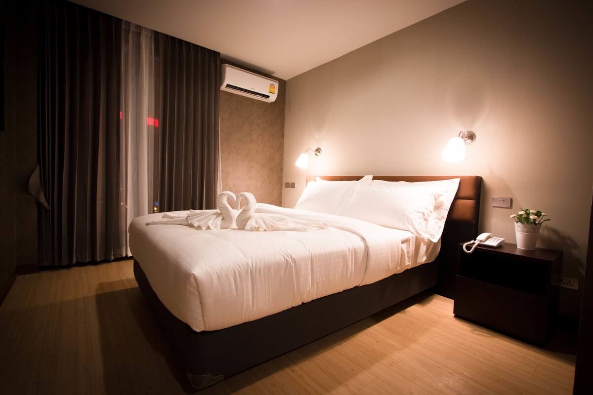 A hotel room with a bed, bedside lamps, a nightstand with a phone and a plant, and an air conditioning unit.