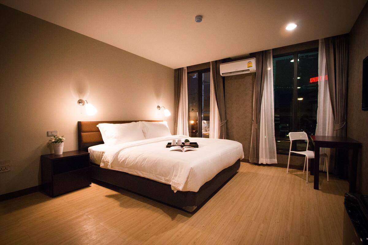 A hotel room with a large bed, bedside lamps, a small table, a chair, and a window with curtains.