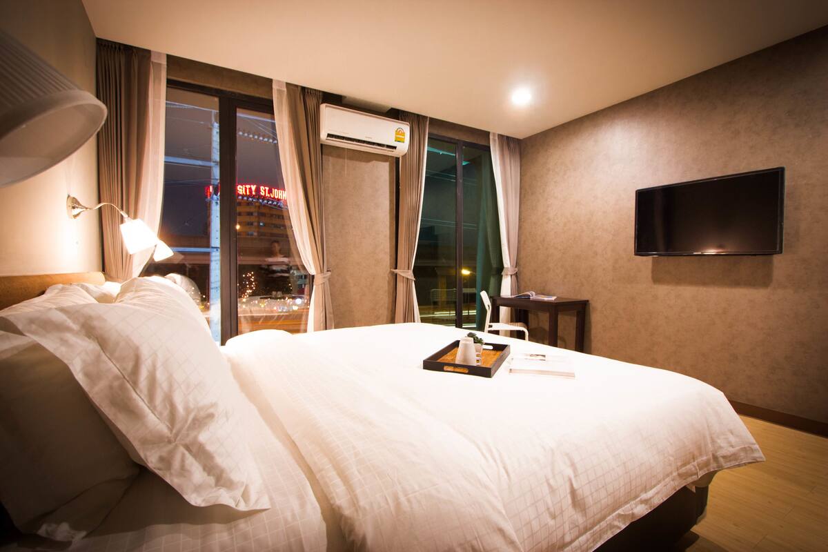 A hotel room with a large bed, an air conditioning unit, a flat-screen TV, and a window with curtains.