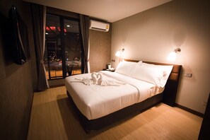 Un cuarto de hotel con una cama grande, una mesita de noche, un aire acondicionado de pared y vista a la ciudad de noche.