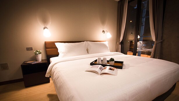 Ein ordentlich bezogenes Bett mit weißer Decke und Kissen, ein Nachttisch mit einer Pflanze und eine Zeitschrift auf dem Bett.