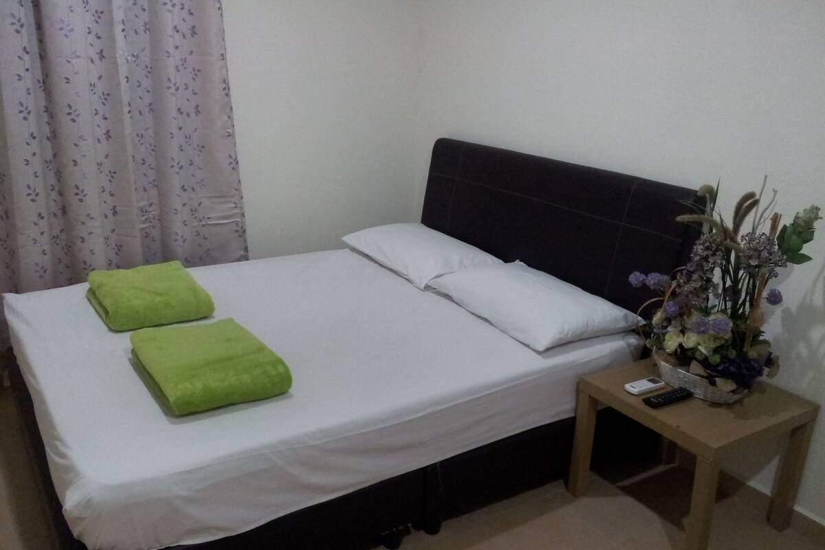 A bed with white sheets and two green towels, a wooden nightstand with a remote control, and a floral arrangement on the nightstand.
