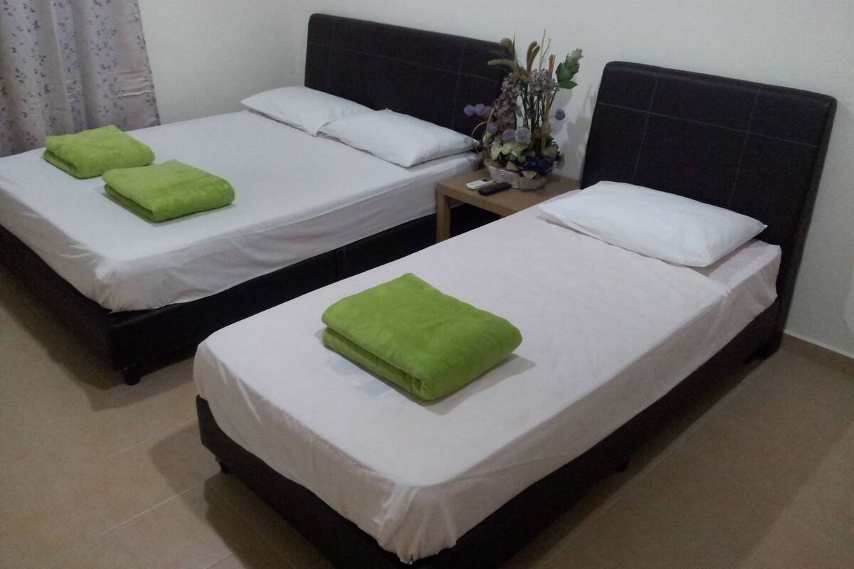 Two single beds with white linens and green towels, a nightstand with a vase of flowers, and a plain wall with a curtain.