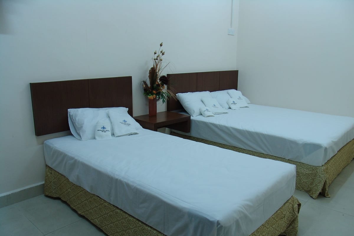 Two single beds with white linens, a wooden headboard, a nightstand with a vase of flowers, and a tiled floor.
