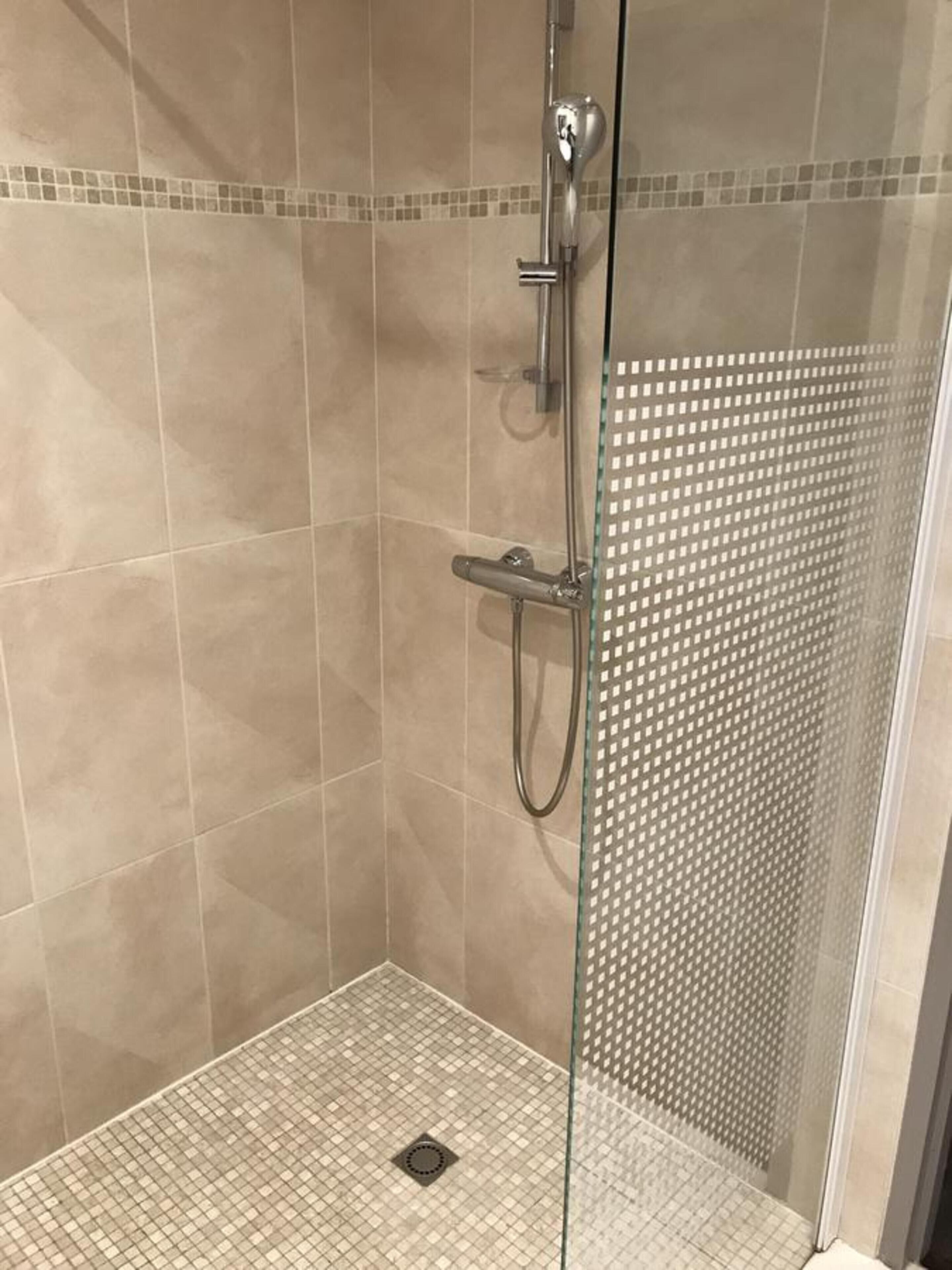 Une douche fermée par des parois en verre, avec des carreaux de couleur beige et un petit drain au centre.