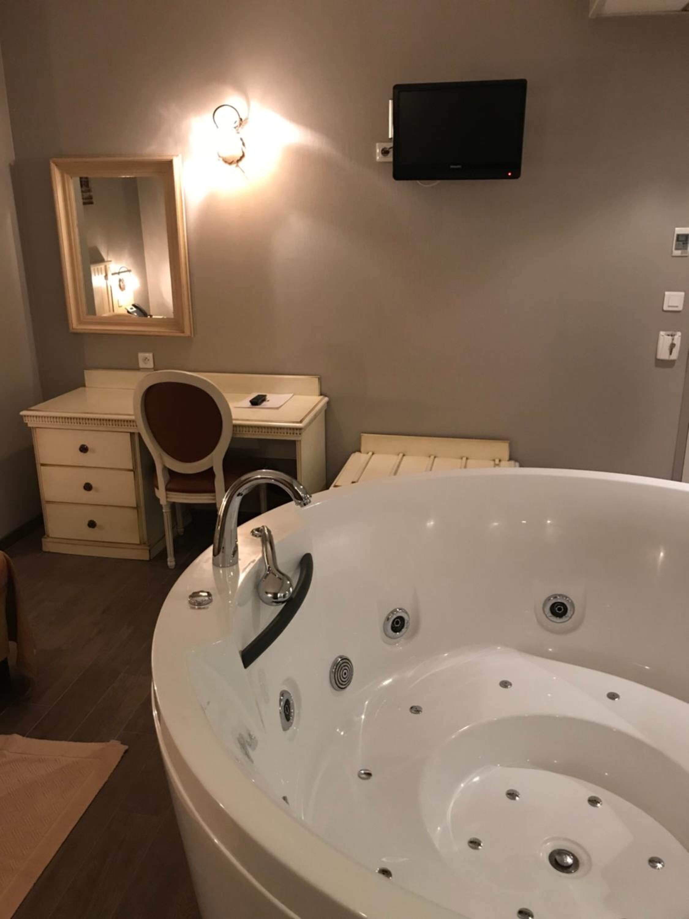 Un baño con una bañera blanca grande, un tocador con espejo, un escritorio y un televisor montado en la pared.
