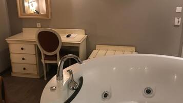 A bathroom with a large white bathtub, a vanity with a mirror, a desk, and a wall-mounted TV.