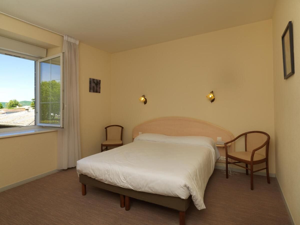 Une chambre à coucher comprenant un lit, deux chaises, une fenêtre avec des rideaux et un luminaire fixé au mur.
