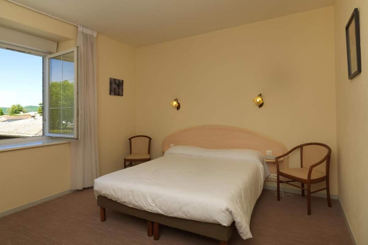 Ein Schlafzimmer mit einem Bett, zwei Stühlen, einem Fenster mit Vorhängen und einer Wandleuchte.