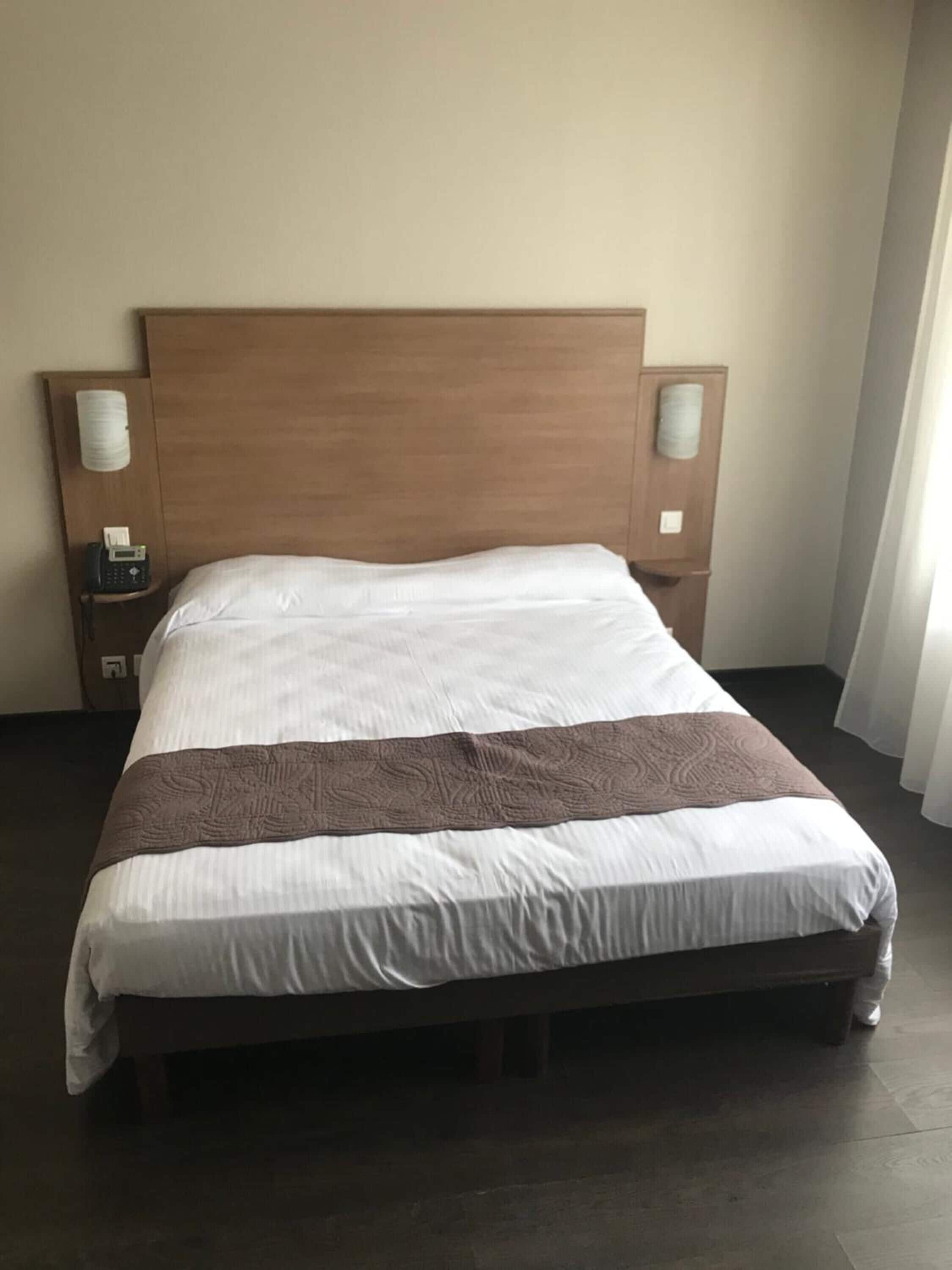 Een standaard hotelkamer met een houten hoofdeinde, een enkel bed met wit beddengoed en een bruine accentkleur, twee nachtkastjes met lampen, en een effen muur.
