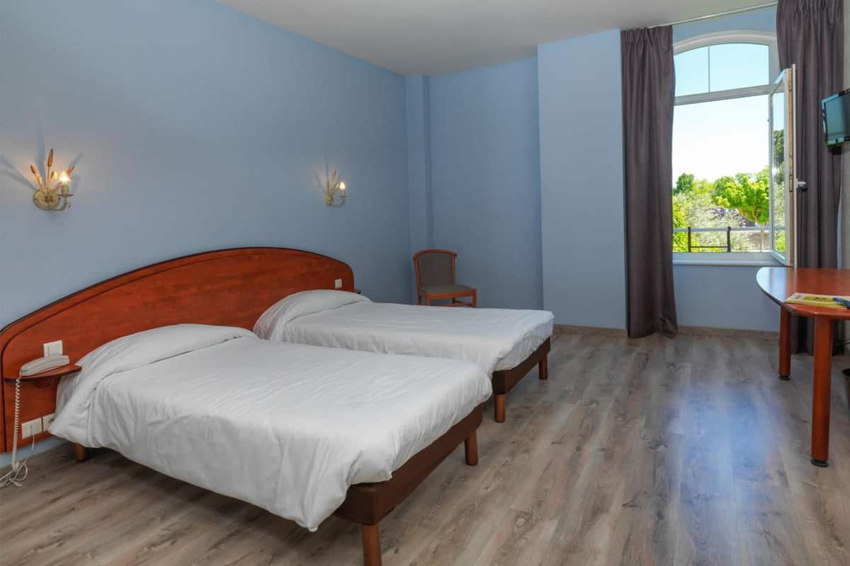 Ein Schlafzimmer mit zwei Betten, einem hölzernen Nachttisch, einem Stuhl, einem Tisch und einem Fenster mit Vorhängen.