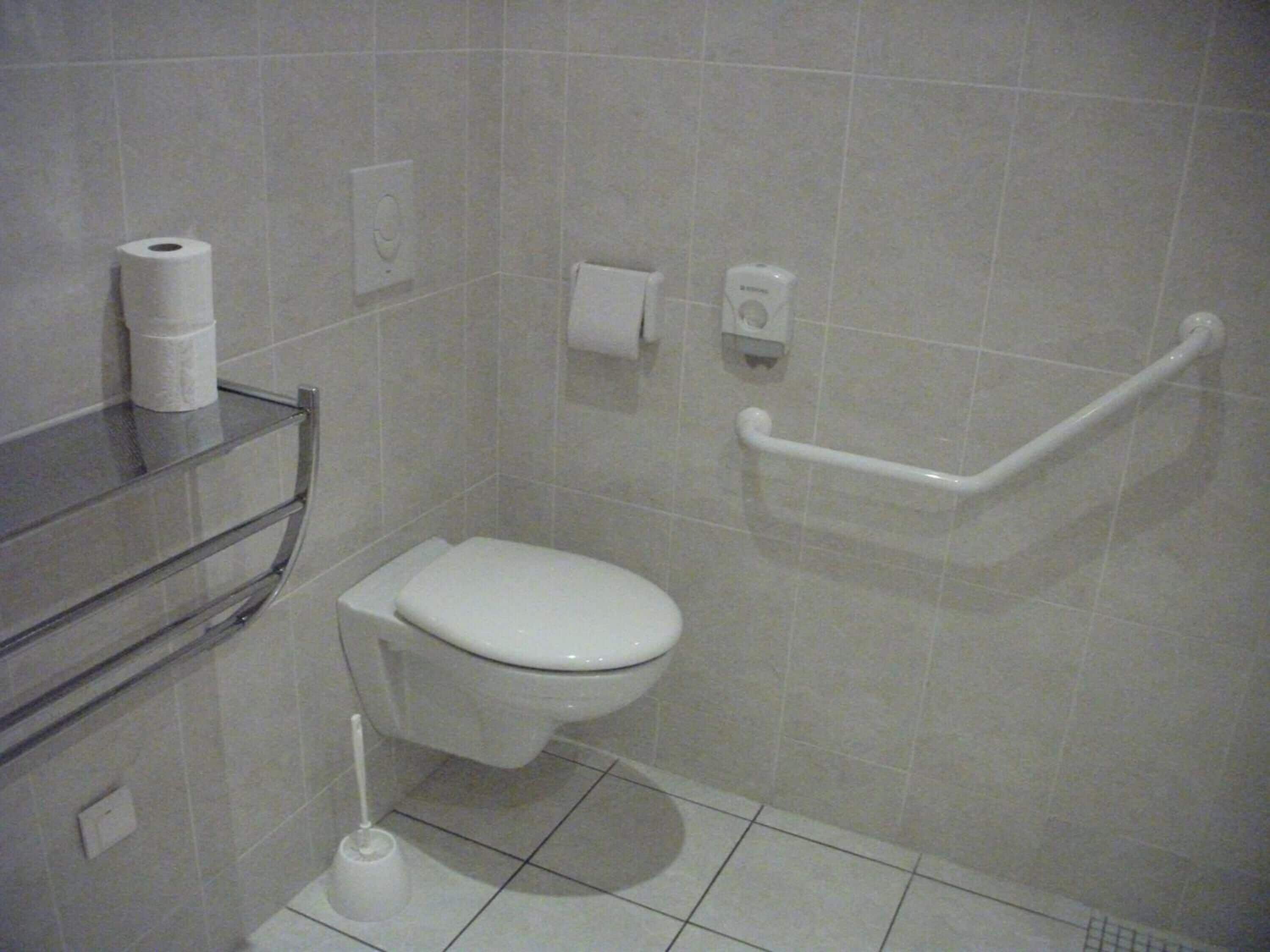 Ein Badezimmer mit Toilette, Handtrockner, Papierhandtuchspender und einer Haltestange.