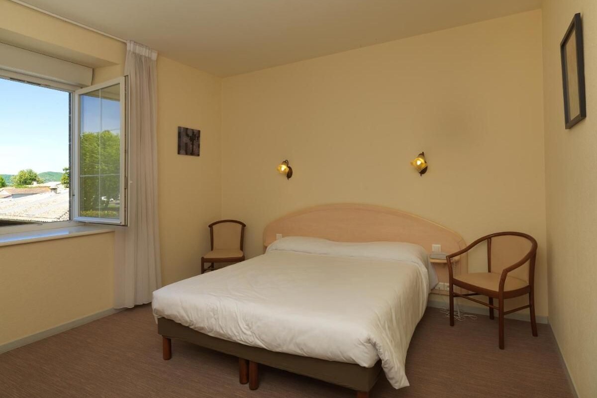 Une chambre à coucher comprenant un lit, deux chaises, une fenêtre avec des rideaux et un luminaire fixé au mur.