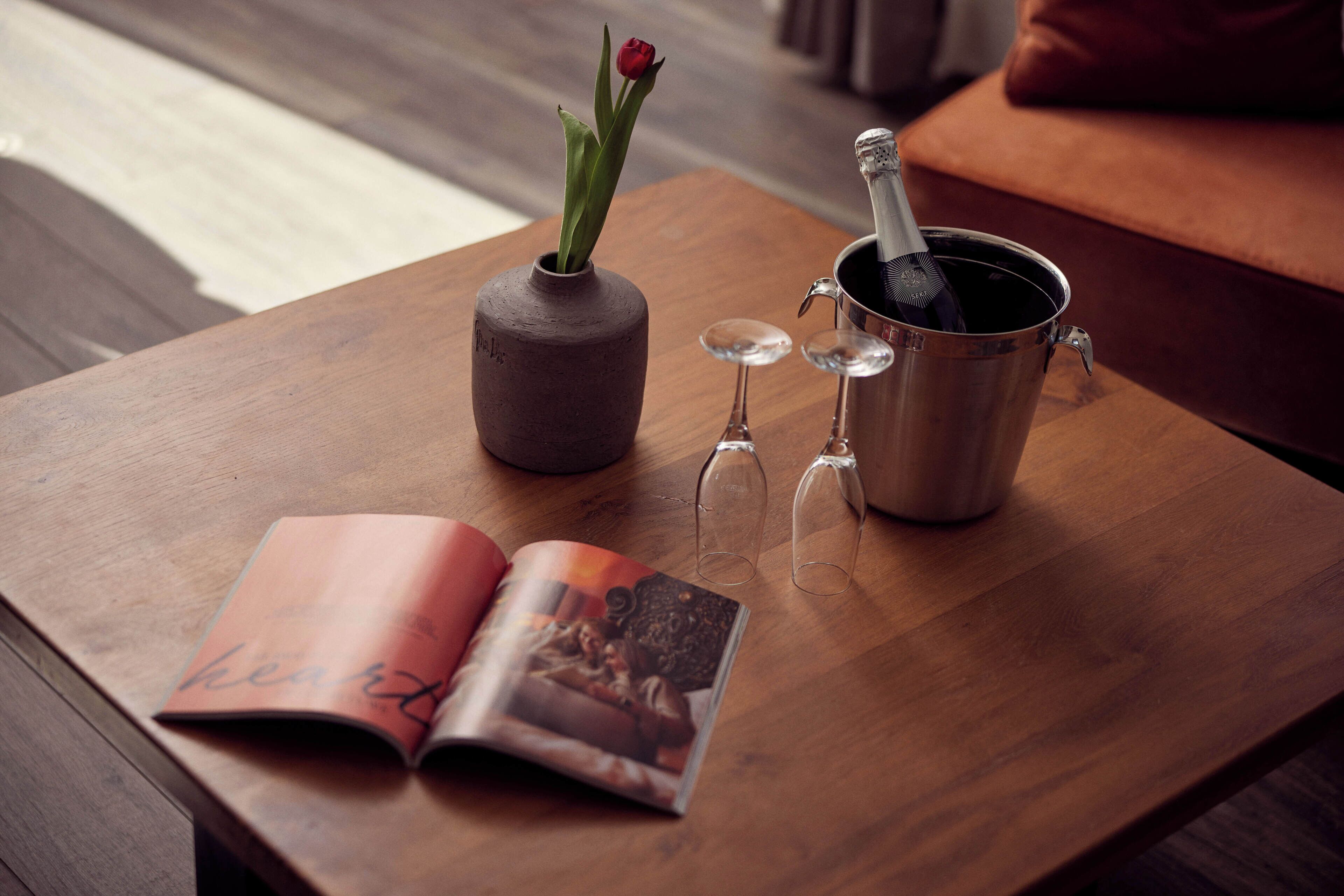 A wooden table with a book, a vase with a flower, a champagne bottle, and two glasses.
