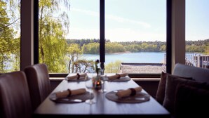 Mesa de comedor lista con platos, cubiertos y un florero, con vista a un lago y un bosque.