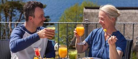 Dos personas cenando al aire libre, con una mesa servida con comida y bebidas, con vista a un cuerpo de agua.