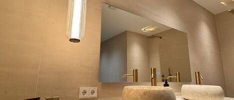 A modern bathroom with a large mirror, two sinks with gold faucets, and a pendant light.