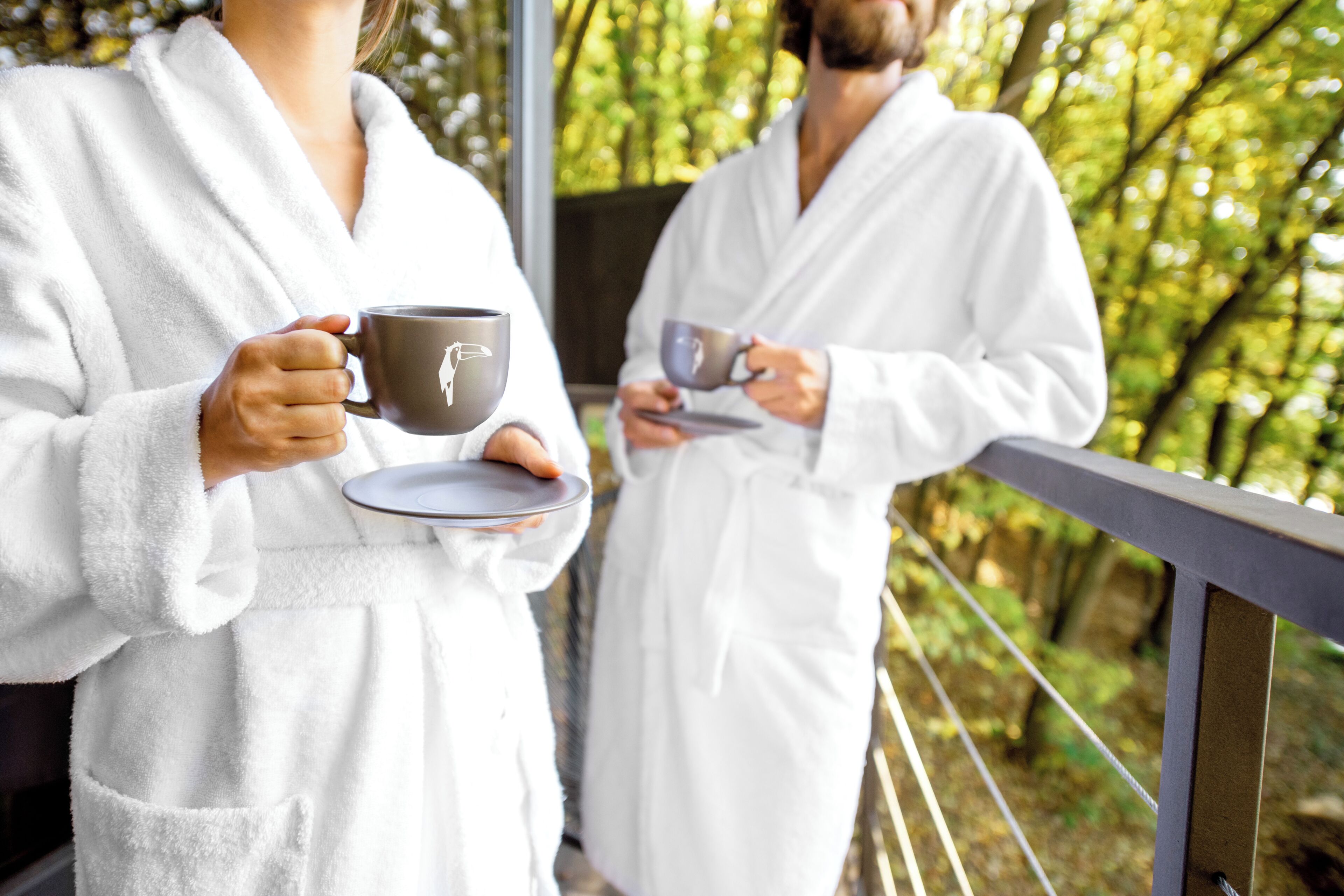 Two individuals in white bathrobes holding cups, standing on a balcony with a railing, surrounded by greenery.