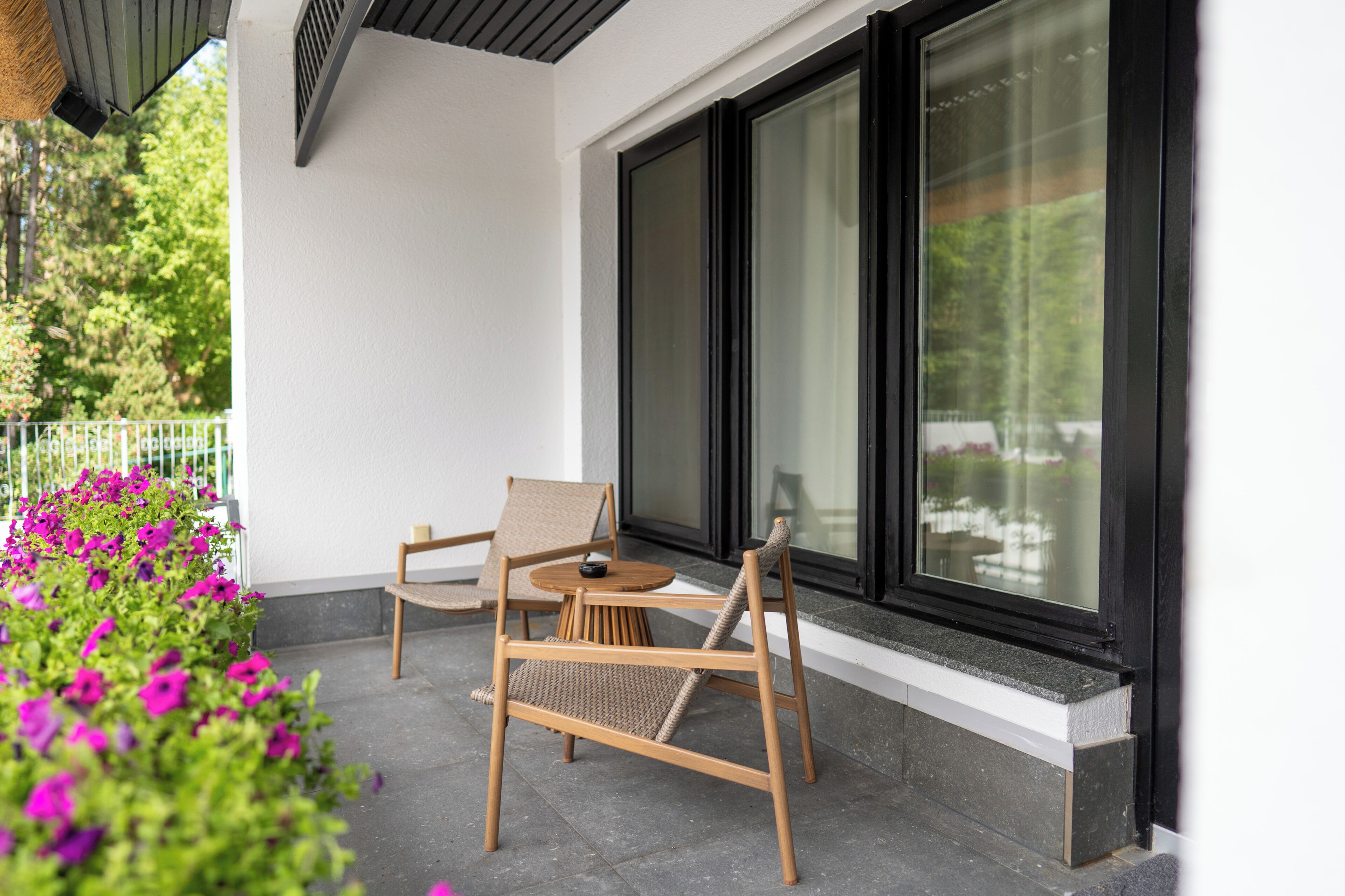 Un patio con dos sillas de madera y una mesita, un ventanal grande y un jardín con flores.