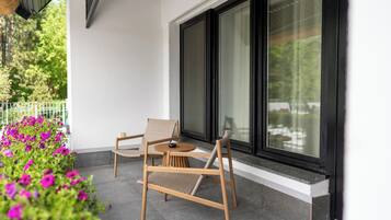 Un patio con dos sillas de madera y una mesita, un ventanal grande y un jardín con flores.