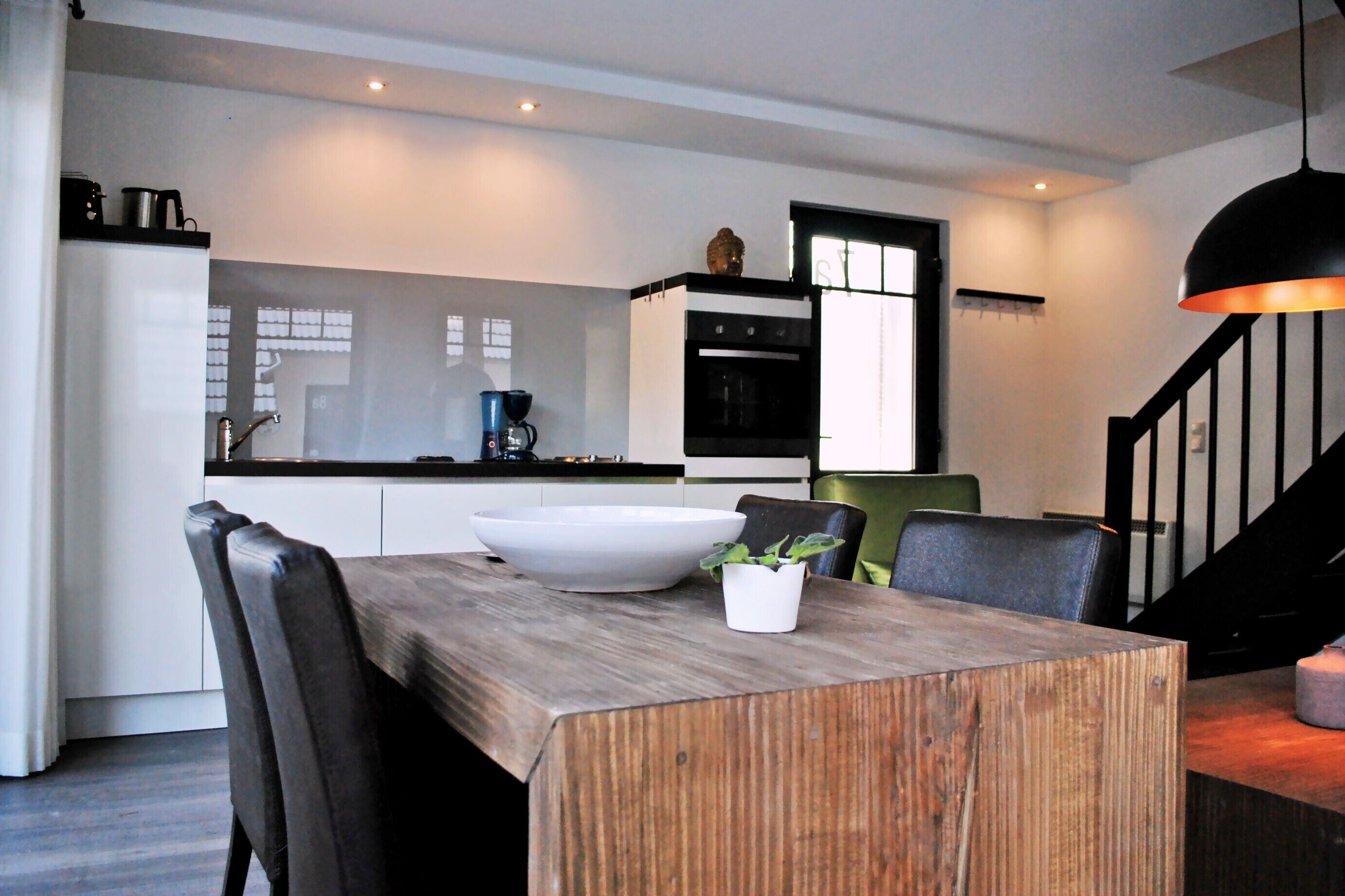 A modern kitchen with white cabinets, a black oven, and a wooden dining table with a white bowl and a potted plant.