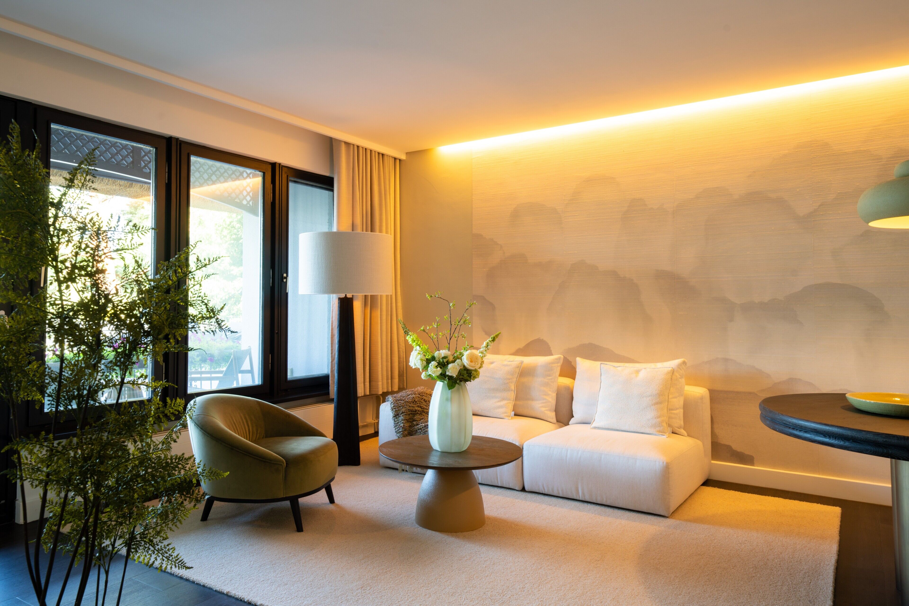 A modern hotel room with a sofa, armchair, round coffee table, and a vase of flowers.