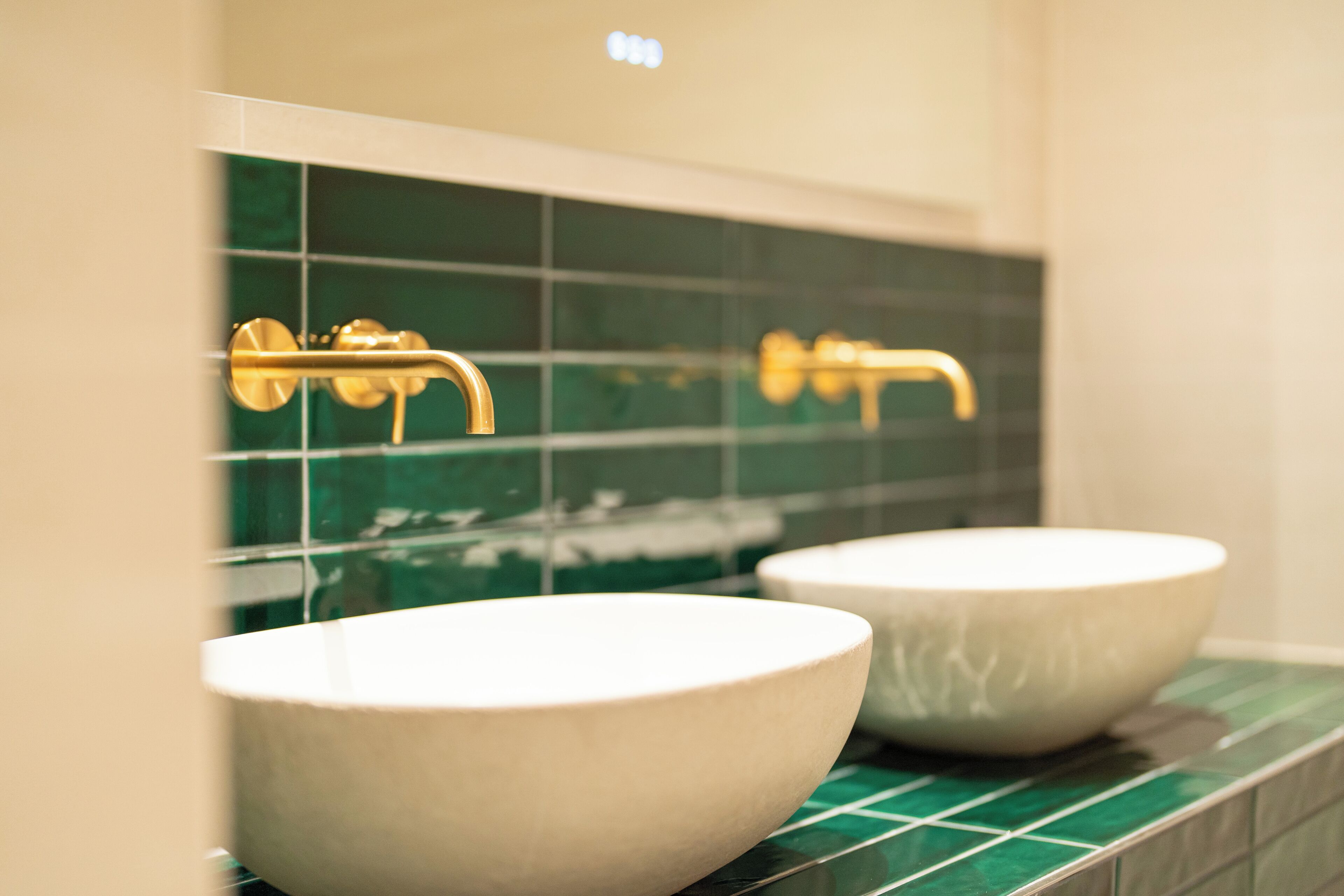 A bathroom with two white sinks, a green tiled backsplash, and gold faucets.