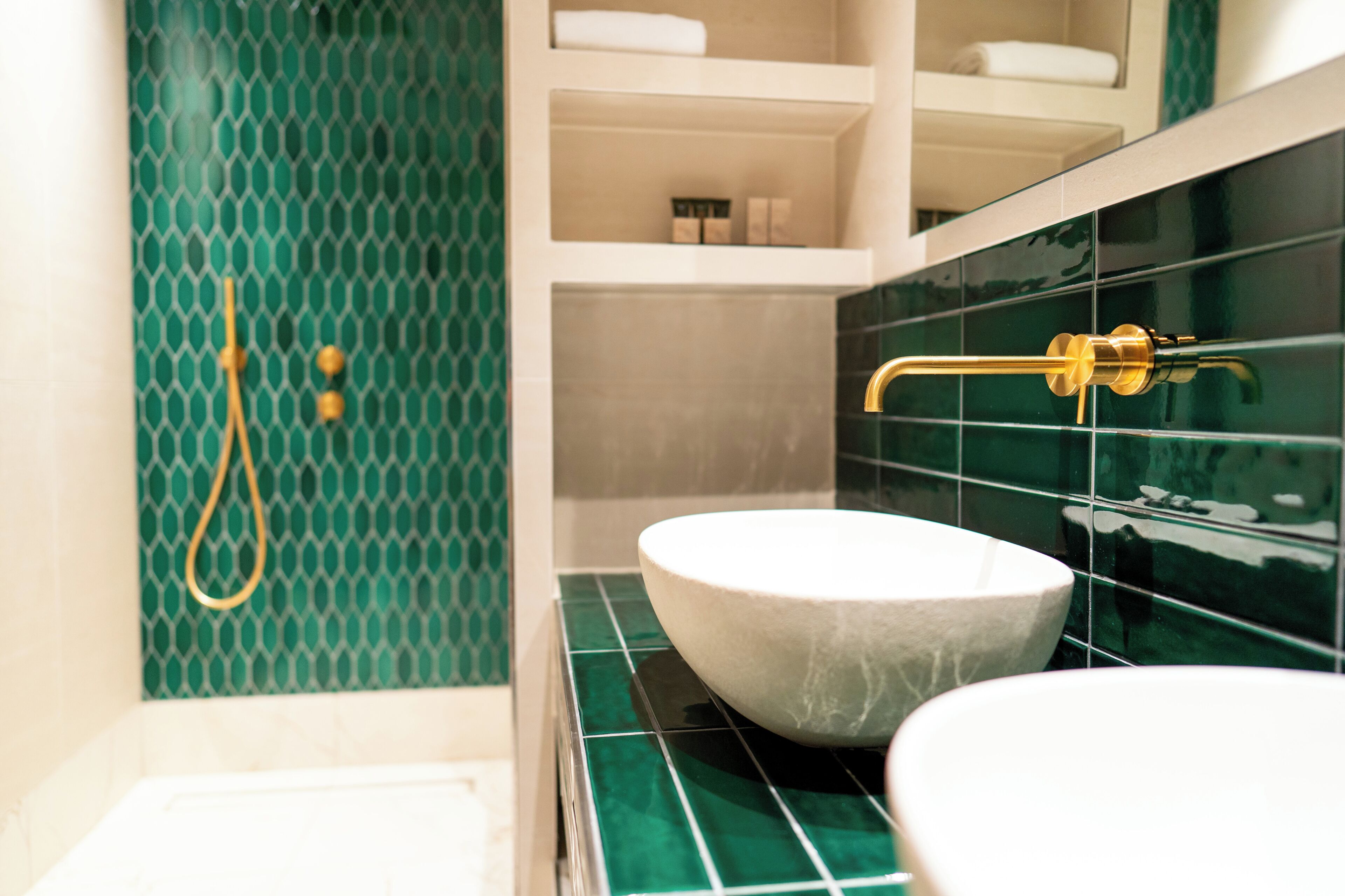 Un baño con un lavamanos blanco, una ducha con detalles dorados y azulejos estampados verdes.