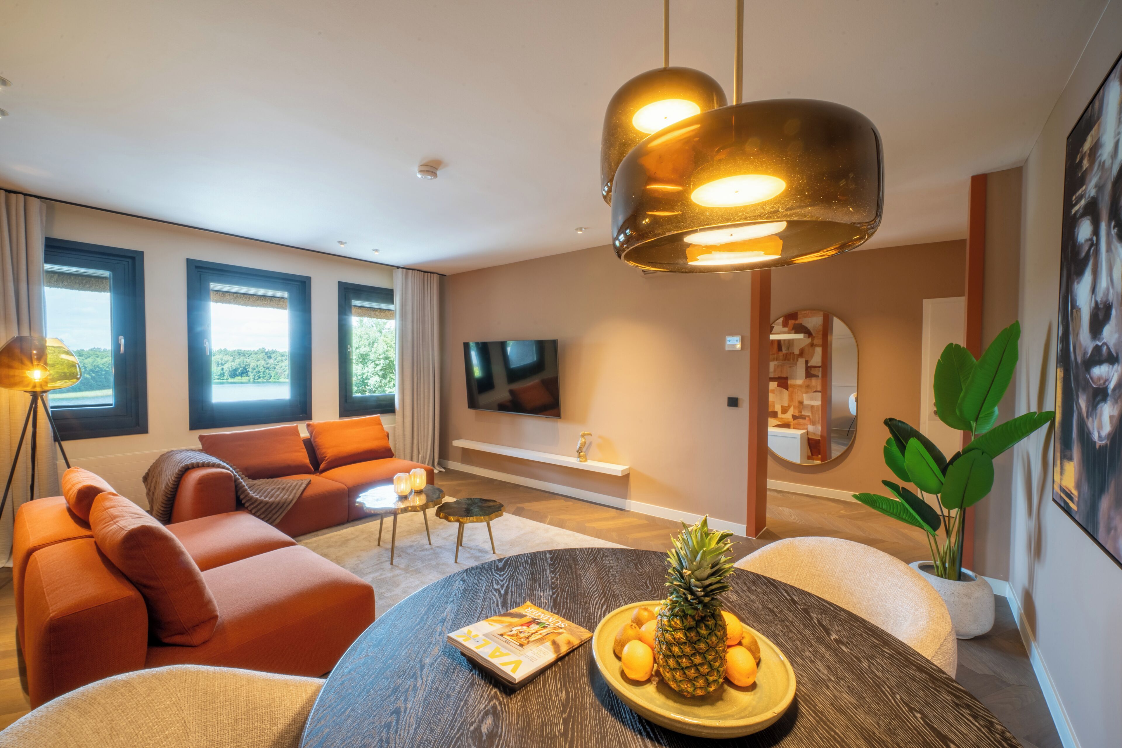 A modern living room with a sofa, a round table with a pineapple and oranges, a TV, and a large window.
