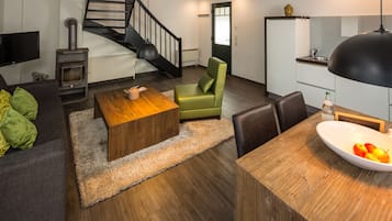 Una sala de estar moderna con un sofá, una mesa de centro de madera, un sillón verde, una mesa de comedor con un cuenco de fruta, una chimenea, una escalera y una zona de cocina.