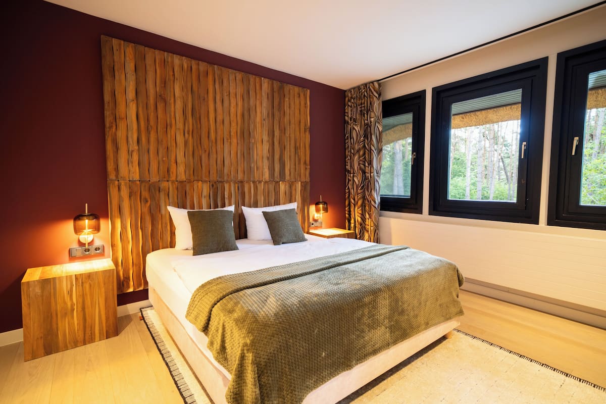 Quarto com cama grande, cabeceira de madeira, luminárias laterais e vista para árvores pelas janelas.