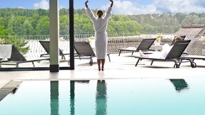 Una persona vestida con una bata blanca, junto a una piscina con vistas a una colina boscosa.