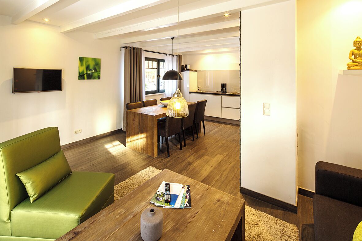 A modern living room with a green sofa, wooden coffee table, and a dining area with a wooden table and chairs.