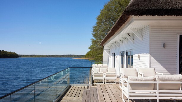 Una terraza de madera con barandas de vidrio que da a una masa de agua, muebles blancos para exteriores y un edificio con techo de paja.