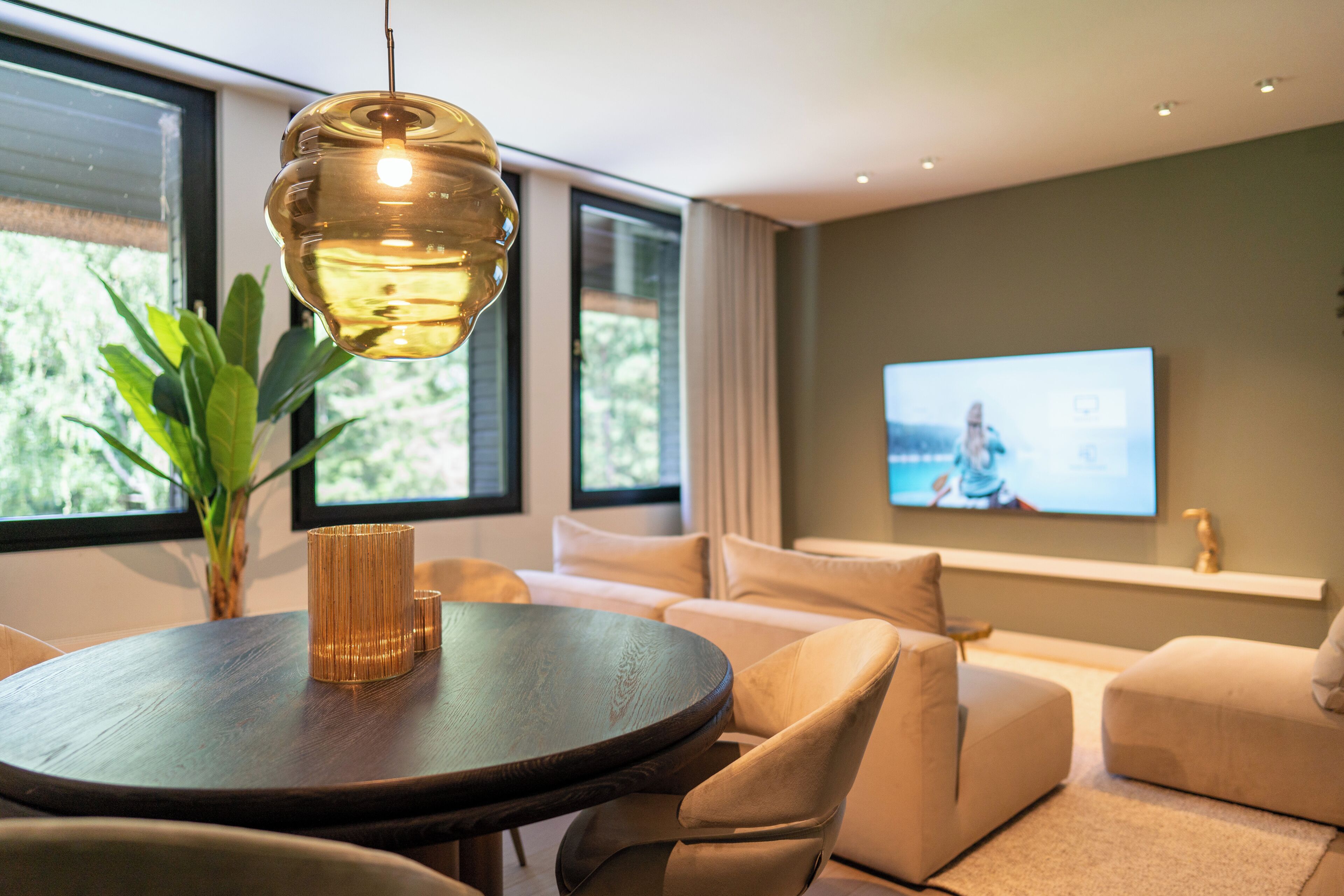 Una sala de estar moderna con una mesa redonda, sofás color beige, una pantalla de televisión grande y una lámpara colgante.