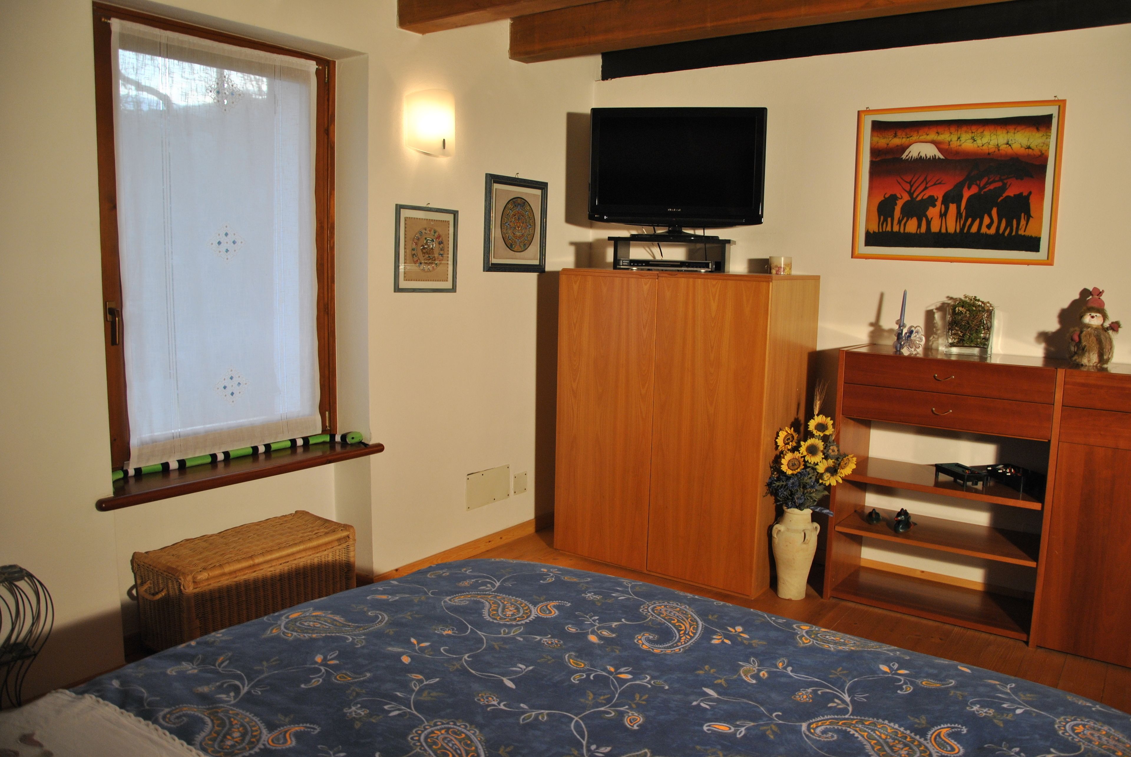A bedroom with a bed, a television, a wooden wardrobe, a window with a curtain, and a wall with framed pictures.
