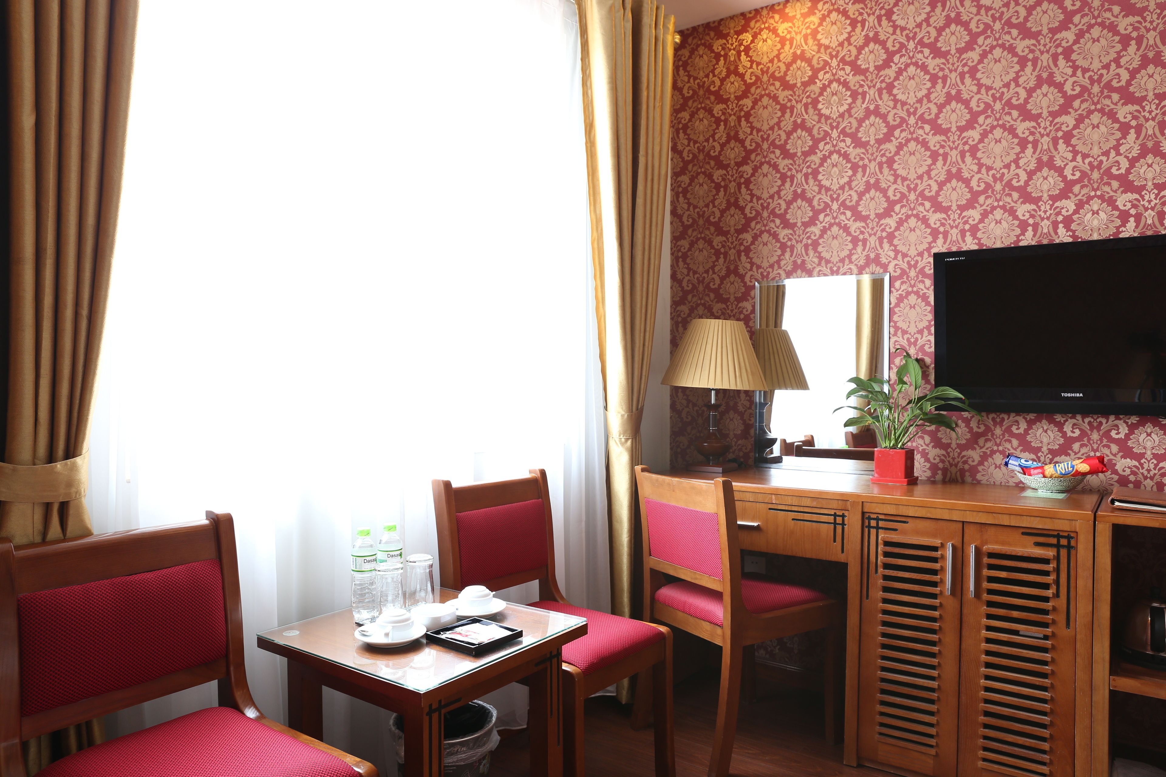 A hotel room with a wooden desk, two chairs, a television, a lamp, and a window with curtains.