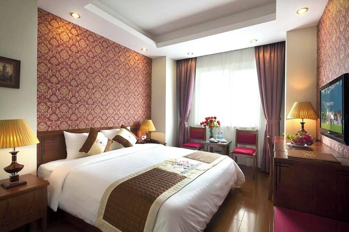 A hotel room with a large bed, two bedside tables with lamps, a TV, a small table with flowers, and a window with curtains.