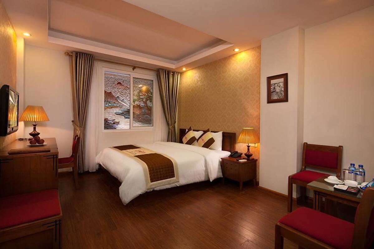 A hotel room with a large bed, bedside tables, a desk with a chair, a window with curtains, and a painting on the wall.
