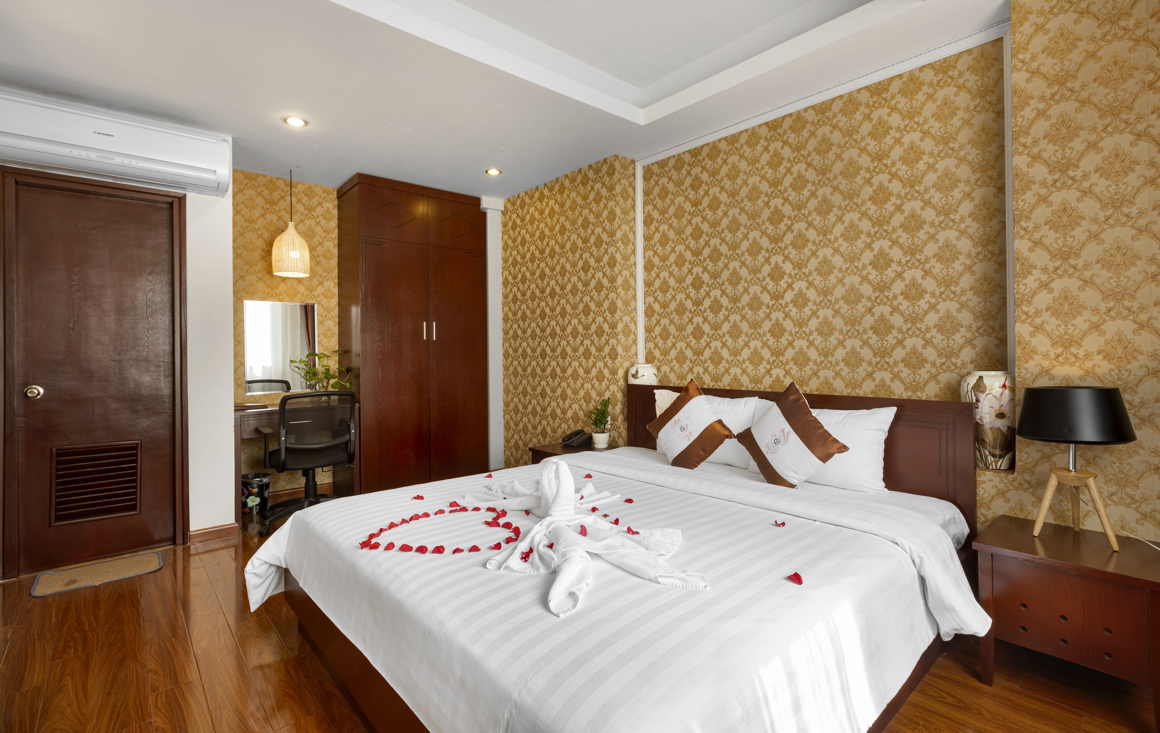 Une chambre d’hôtel avec un grand lit, un plancher en bois, un bureau avec une lampe, une armoire et une salle de bain avec un miroir et un lavabo.