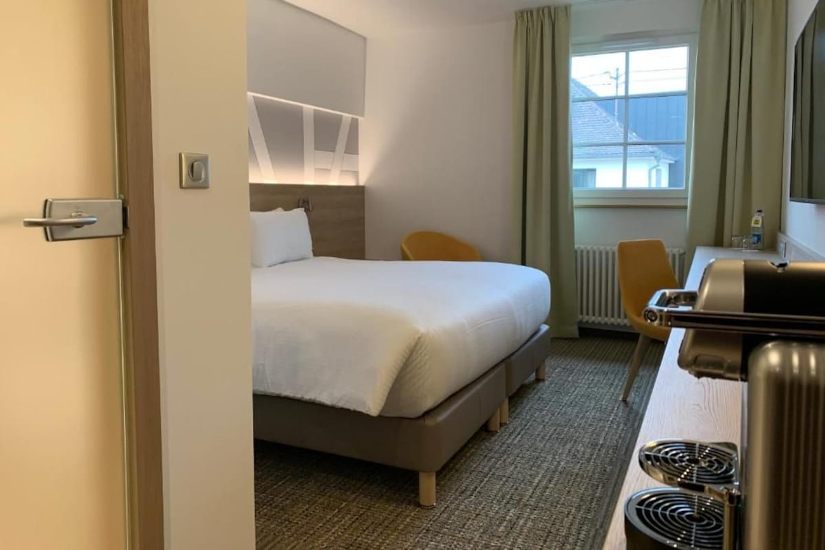 Habitación de hotel con cama, escritorio con silla, radiador y ventana con cortinas.