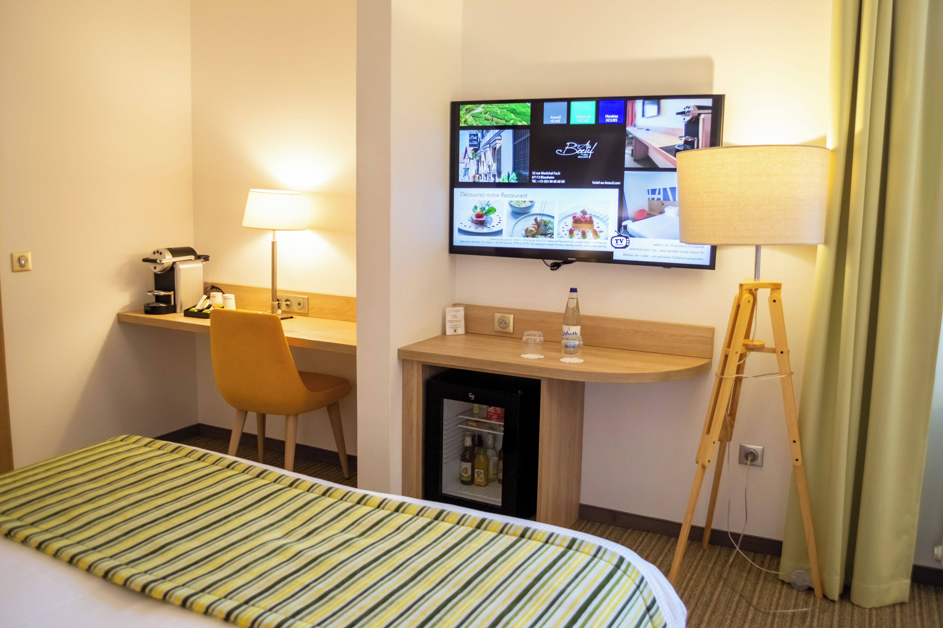 Ein Hotelzimmer mit Bett, Schreibtisch mit Stuhl, Wandfernseher, Lampe und einem kleinen Kühlschrank.