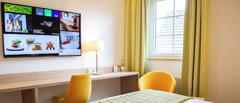 A hotel room with a bed, a desk, two chairs, a TV mounted on the wall, and a window with curtains.