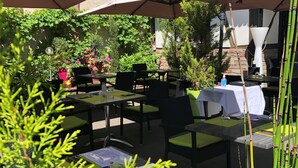 Un espace extérieur aménagé en terrasse, avec des tables et des chaises placées sous de grands parasols, entouré de verdure et de plantes en pot.