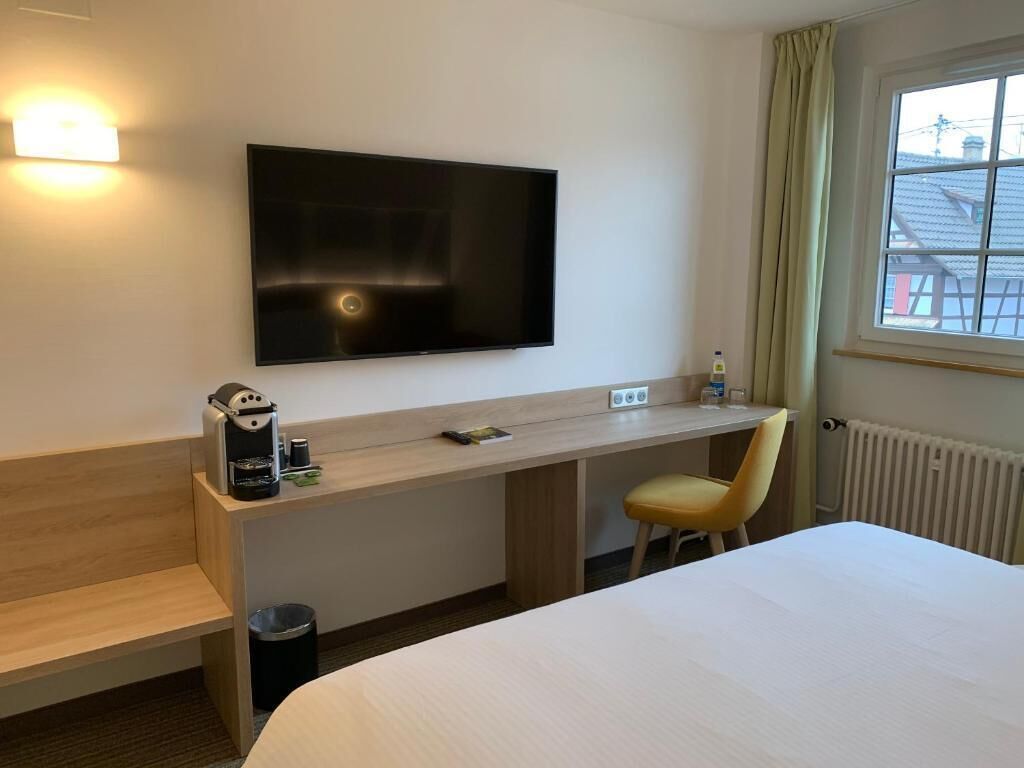 A hotel room with a bed, a desk, a chair, a TV, a coffee machine, and a window with curtains.