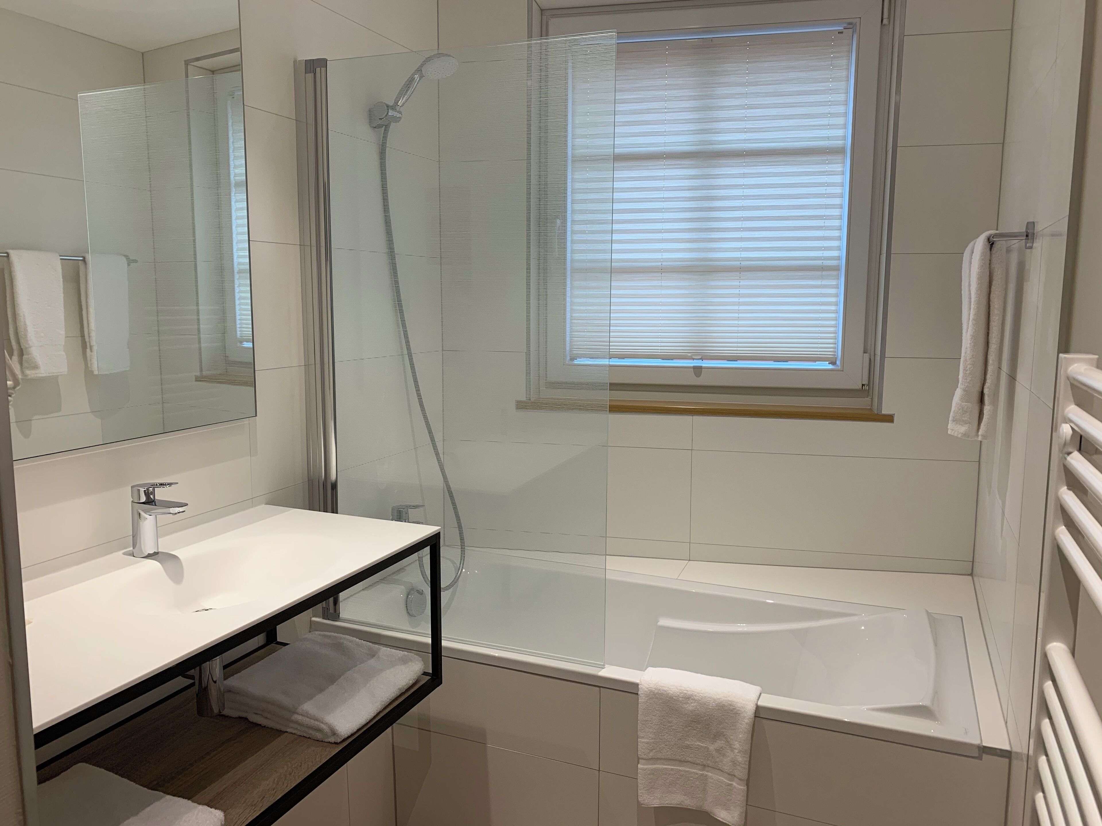 Une salle de bain moderne dotée d’une douche à parois de verre, d’une baignoire, d’un meuble-lavabo et d’une fenêtre avec des stores.
