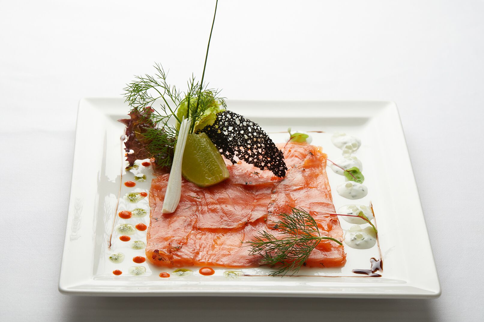 A square white plate with a neatly arranged dish of salmon, garnished with dill, a black edible flower, and a slice of lime.