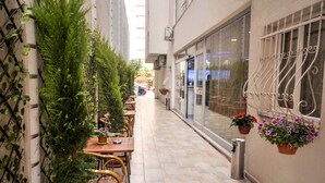 Un café al aire libre con mesas y sillas, plantas en macetas y un basurero.